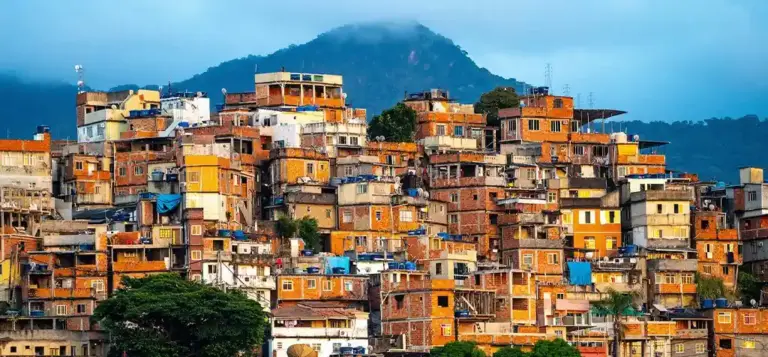 Comunidade de casas autoconstruídas ocupando a encosta de um morro, com montanha ao fundo