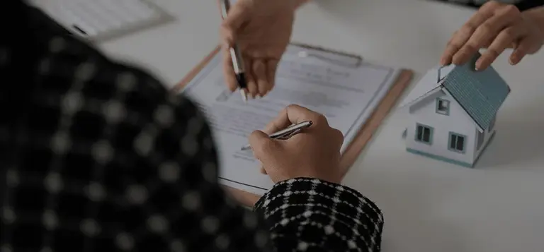 Mãos assinando um documento ao lado de uma maquete de casa
