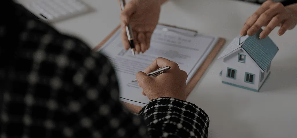 Mãos assinando um documento ao lado de uma maquete de casa