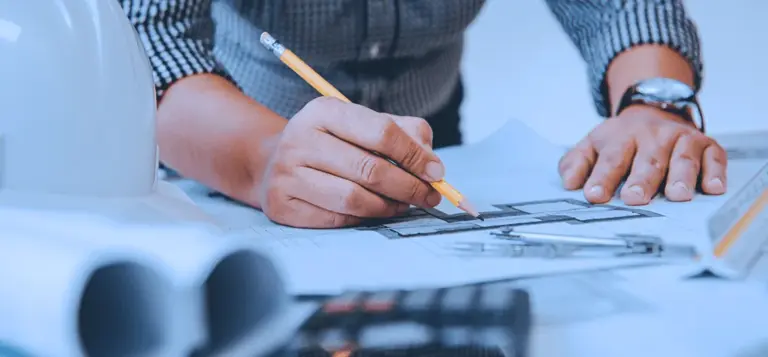Mãos desenhando sobre uma planta técnica ao lado de um capacete de obra